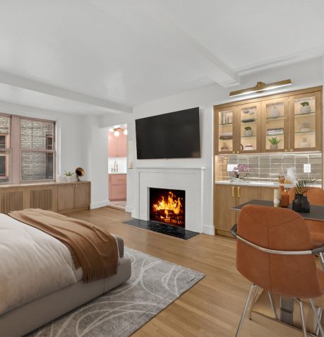Cozy modern bedroom with a lit fireplace, wall-mounted TV, wooden floors, a bed with brown throw, and a small dining area with orange chairs.