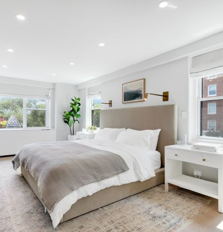 Bright bedroom with large beige upholstered bed, gray blanket, white bedding, white nightstands, and windows letting in natural light.