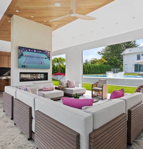 Modern outdoor patio with grey sectional sofa, pink cushions, ceiling fan, mounted TV showing tennis match, and view of lawn and swimming pool.
