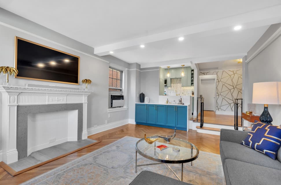 Modern living room with gray walls, white fireplace, mounted TV, blue cabinet, glass coffee table, and gray couch with blue and gold pillows.