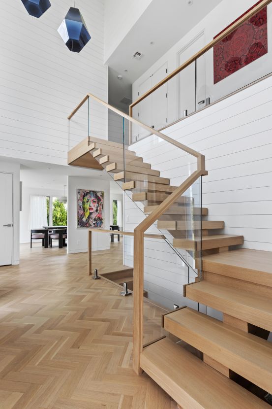 Modern wooden staircase with glass railing in a bright, open interior featuring herringbone wood floors and colorful artwork on the wall.