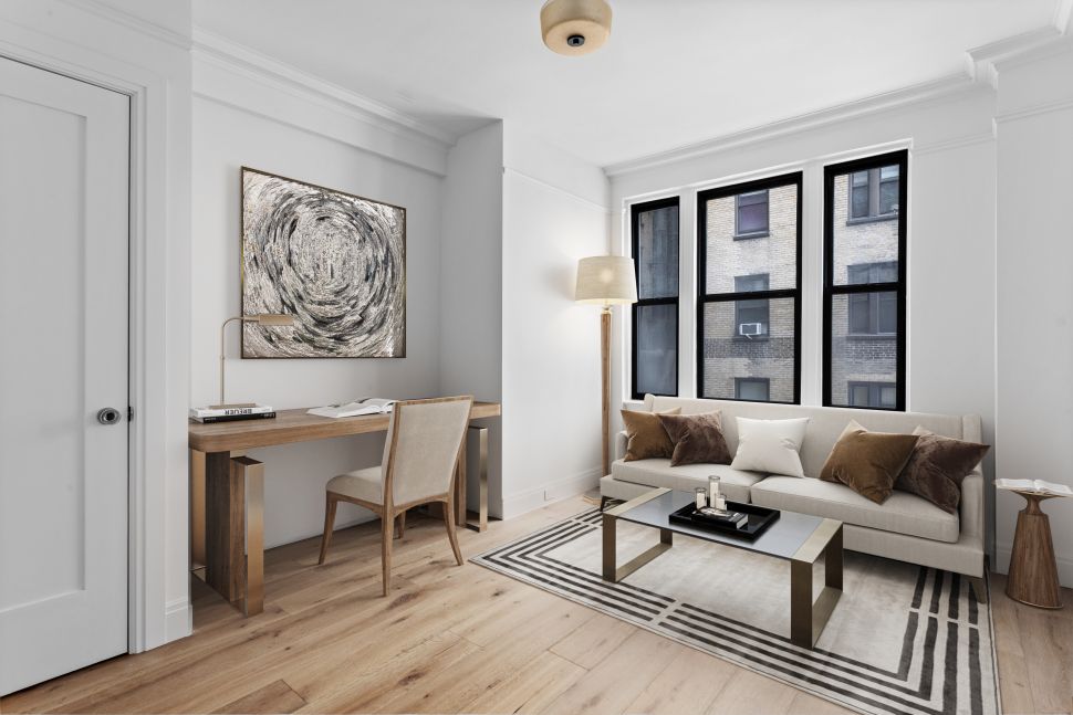 Modern living room with a beige sofa, striped area rug, glass-top coffee table, wooden desk with chair, abstract wall art, and large windows.