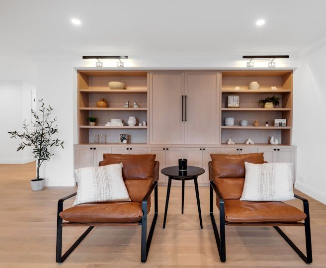 Modern living room with two brown leather chairs, a small black round table between them, built-in wooden shelves with decorative items, and a potted plant to the left.