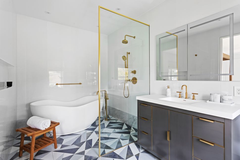 Modern bathroom with white freestanding bathtub, wooden stool with rolled towel, glass shower enclosure with gold fixtures, and dark vanity with gold hardware and mirror above.
