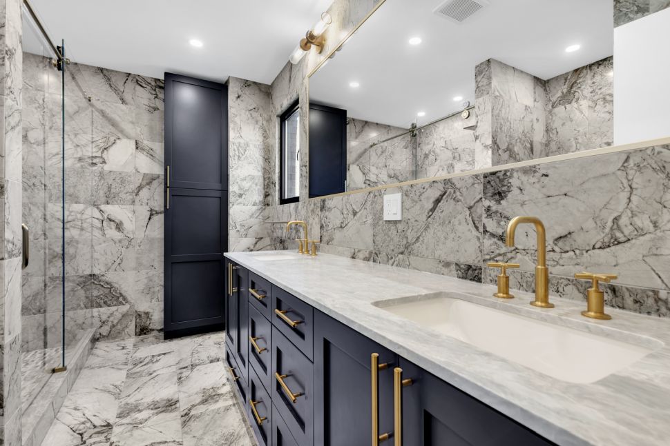 Modern bathroom with marble walls and floor, navy blue cabinetry with gold handles, dual sinks, and a glass-enclosed shower.