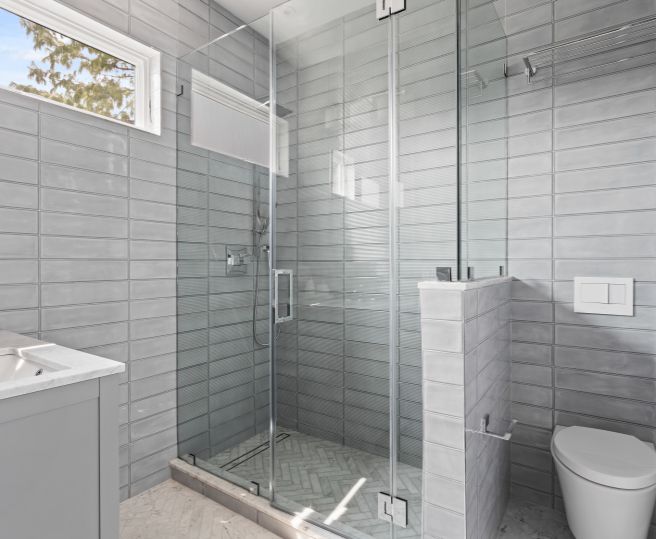 Modern bathroom with gray tiled walls, glass shower enclosure, white sink countertop, and toilet.