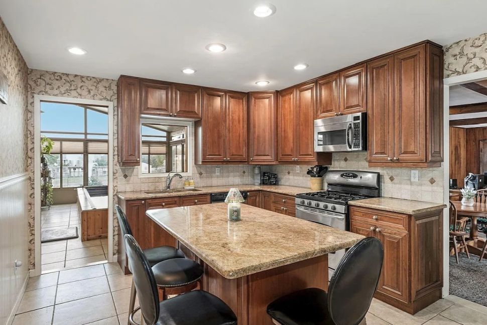 Kitchen with wooden cabinets, granite island with three black chairs, stainless steel stove and microwave, and tiled floor.
