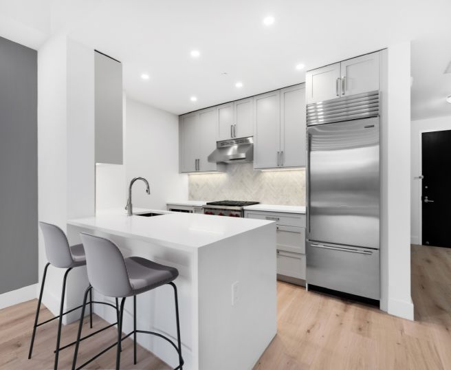 Modern kitchen with gray cabinets, stainless steel appliances, white island with two gray bar stools, and light wood flooring.