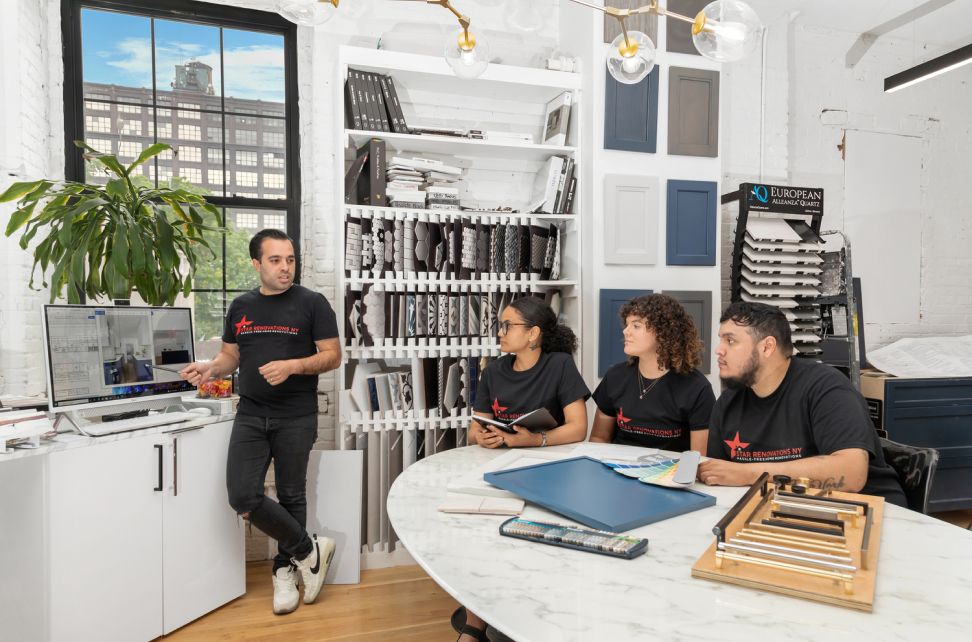 A man presents design plans on a desktop monitor to three seated colleagues wearing matching black T-shirts in a bright office with samples on the walls and shelves.