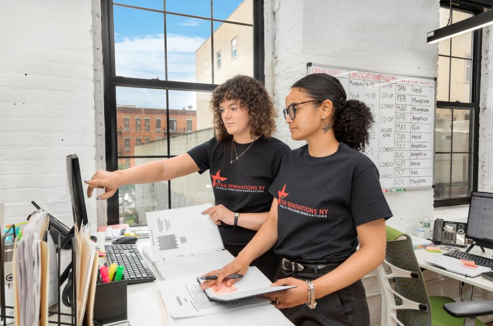 Two women in black Star Renovations NY shirts working together at a desk with documents and a computer in a bright office.