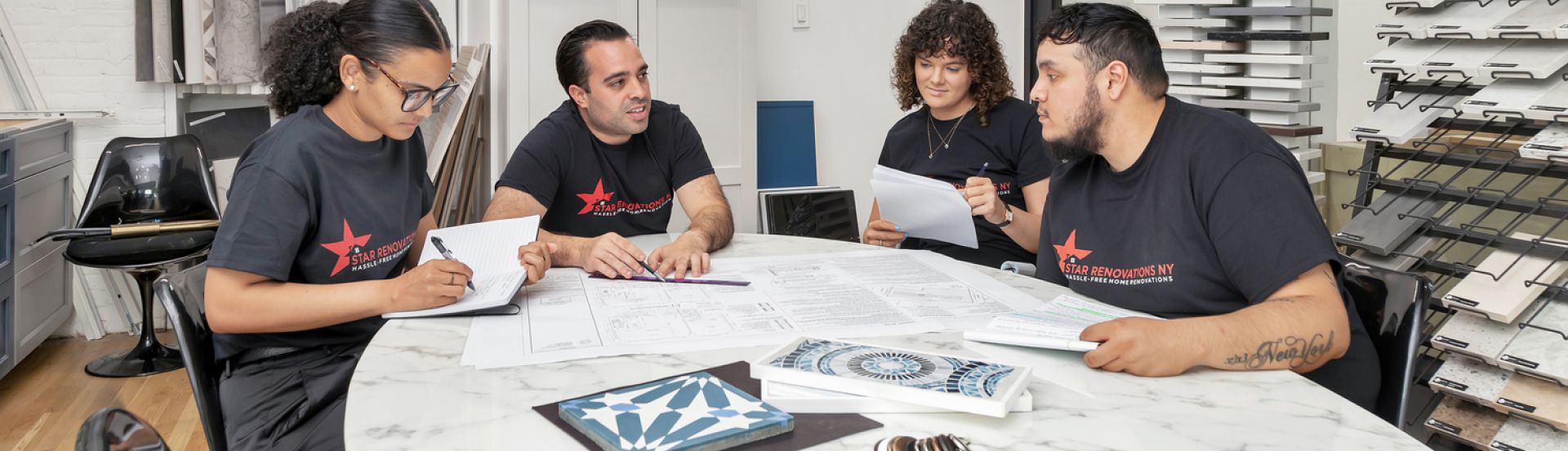 Four people in black Star Renovations NY shirts review blueprints and tiles at a marble table in a design showroom.
