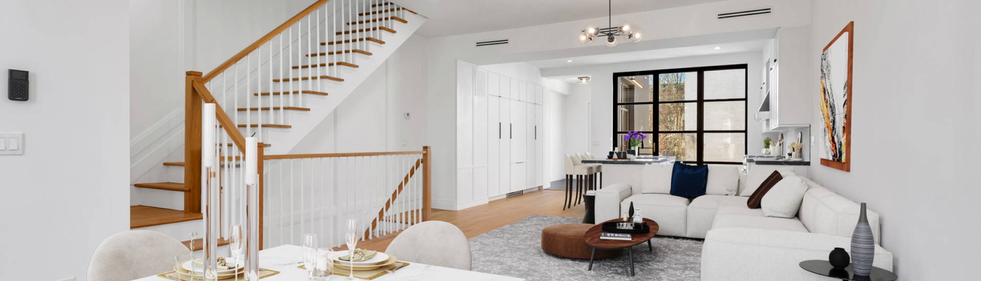 Modern open-plan living and dining area with white walls, wooden staircase, gray sofa, and large windows.