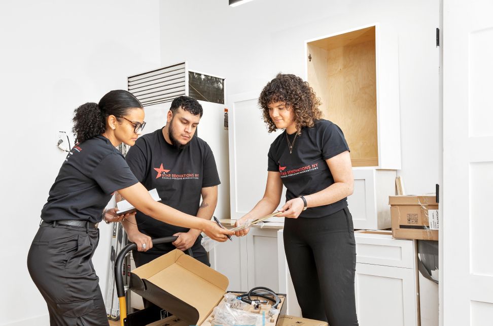 Three people in black Star Renovations NY shirts discussing papers inside a kitchen under renovation.