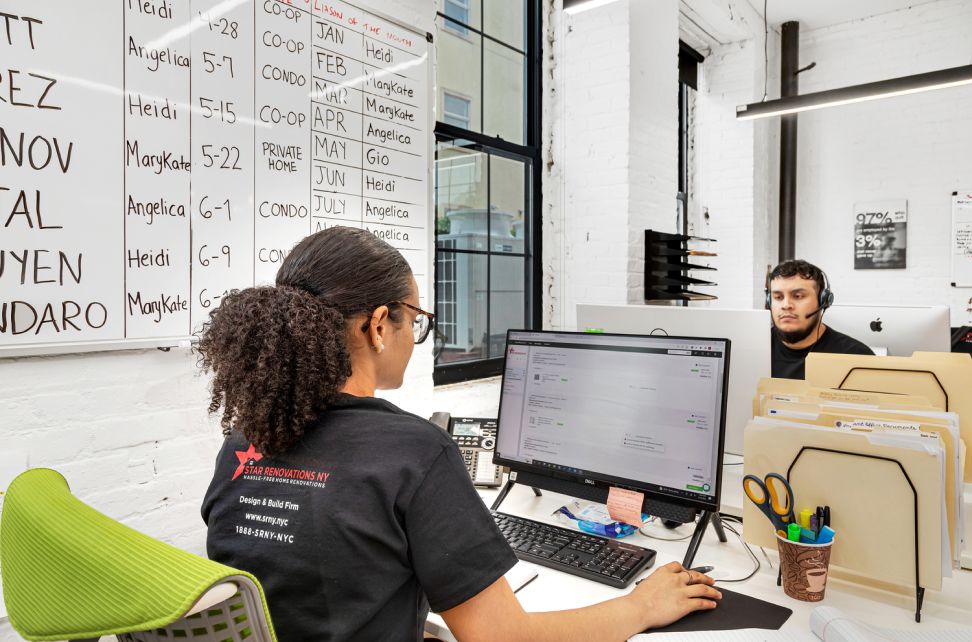 Two office workers at desks facing computers, with a whiteboard listing names and dates on the wall.