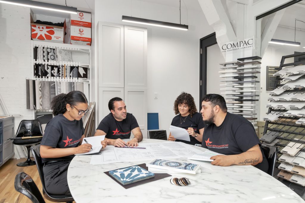 Four people in matching black Star Renovations NY t-shirts sitting around a marble table discussing blueprints and tile samples in a showroom.