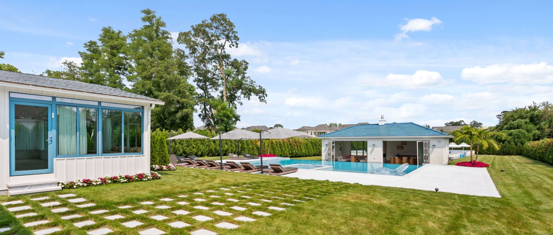 Modern backyard with blue-roofed pool house, infinity pool, lounge chairs under umbrellas, and lush green lawn.