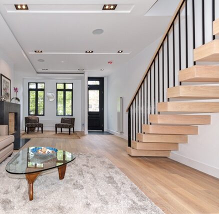 Modern living room with floating wooden staircase, glass coffee table on a beige rug, two chairs by black-framed windows, and a black door.