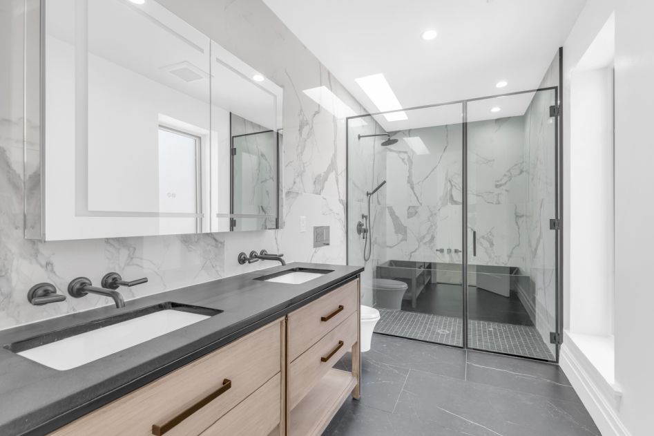 Modern bathroom with dual sinks set in a black countertop, light wood vanity, marble walls, and a glass-enclosed shower.