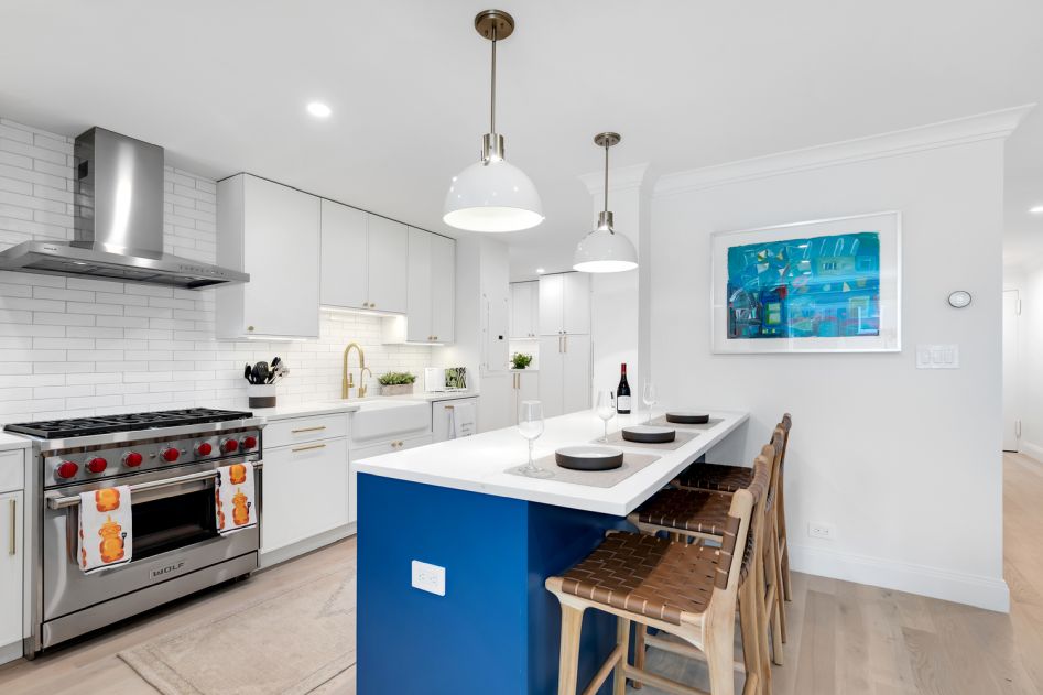 Modern kitchen with a blue island, white countertops, wooden bar stools, stainless steel stove, and white cabinetry.