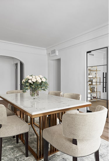 Modern dining room with a white marble table, beige upholstered chairs, and a vase of white flowers as centerpiece.