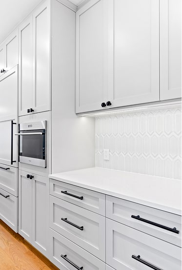Modern white kitchen cabinets with black handles, built-in oven, white geometric tile backsplash, and wooden floor.