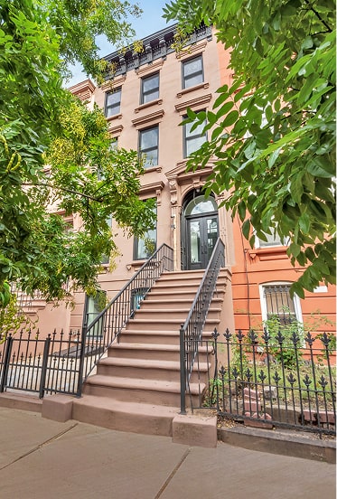Front view of a brownstone building with a tall staircase and black iron railings, framed by green tree branches.