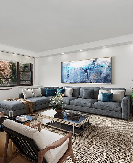 Modern living room with large gray sectional sofa, blue and white cushions, glass coffee table with books and plant, armchair, large abstract blue artwork on white wall, and a window showing outdoor trees.