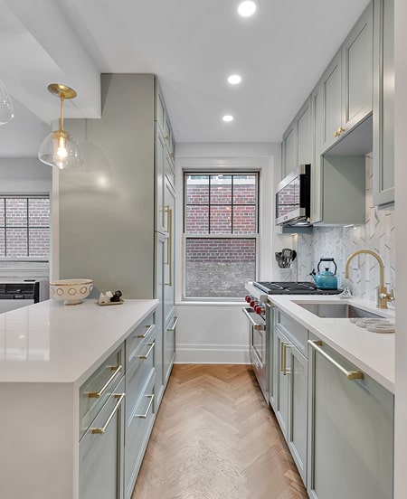 Modern galley kitchen with light green cabinetry, gold handles, white countertops, hardwood herringbone floor, and a window overlooking a brick wall.
