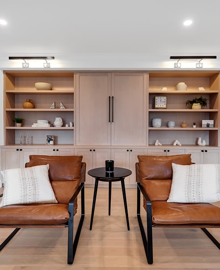 Modern living room with two brown leather chairs, a small black round table between them, and a built-in wooden shelving unit with decorative items in the background.
