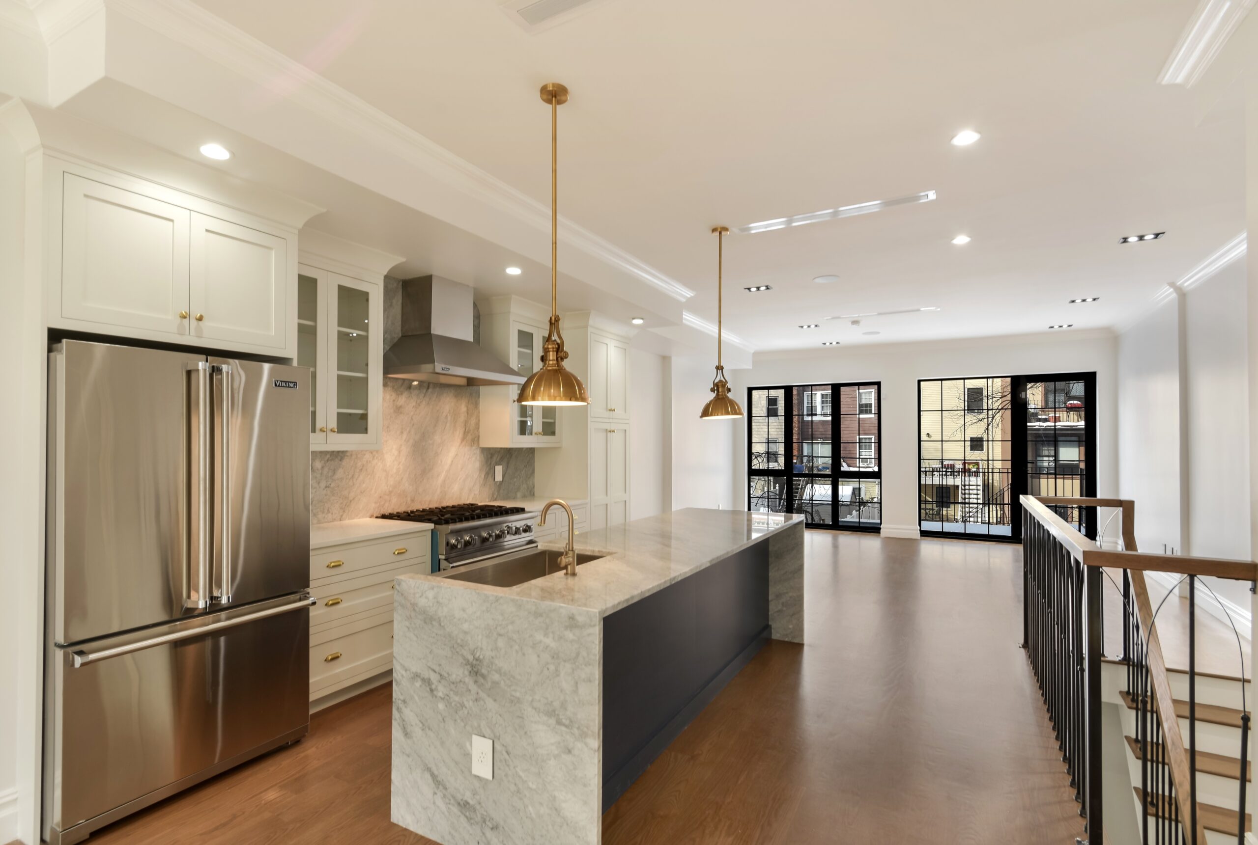 Modern kitchen with marble island, brass pendant lights, stainless steel refrigerator, and large windows overlooking city buildings.