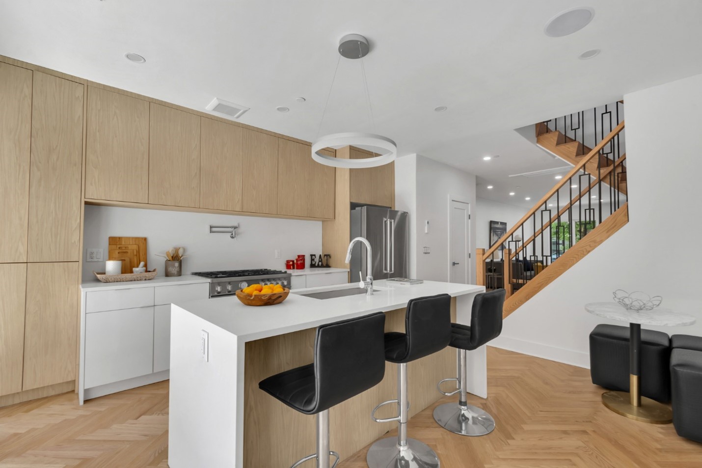 Modern kitchen with light wood cabinets, white island with sink and three black bar stools, stainless steel refrigerator, and wooden staircase with black metal railing.