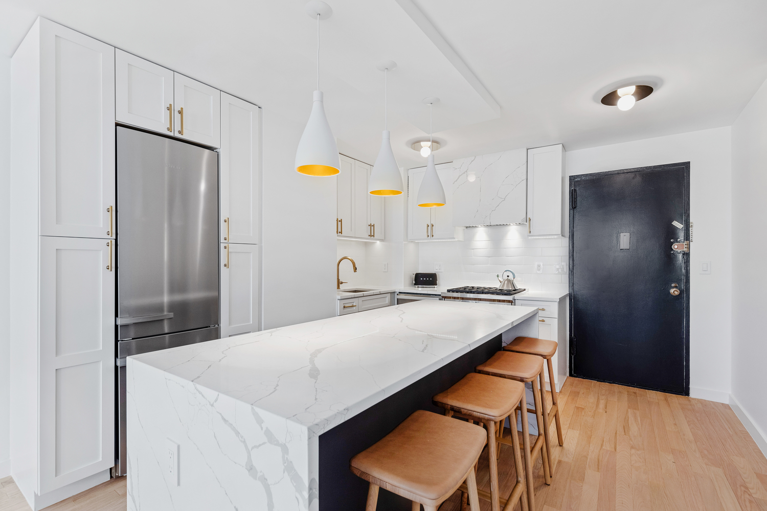 Modern kitchen with white marble island, four brown bar stools, white cabinets, stainless steel refrigerator, and black door.