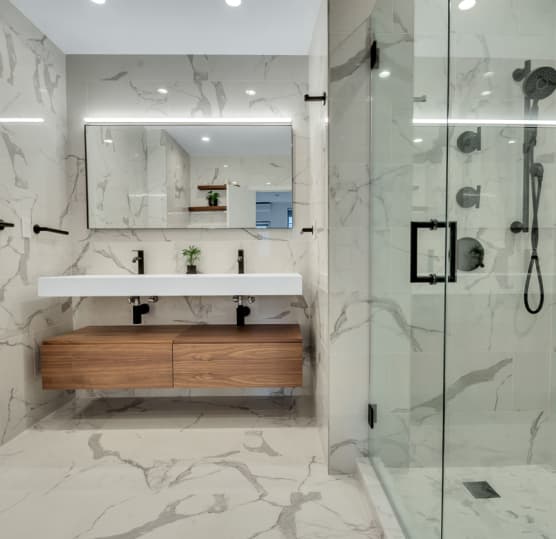 Modern bathroom with white marble walls and floor, a double sink with wooden cabinet, a large mirror, and a glass-enclosed shower with black fixtures.