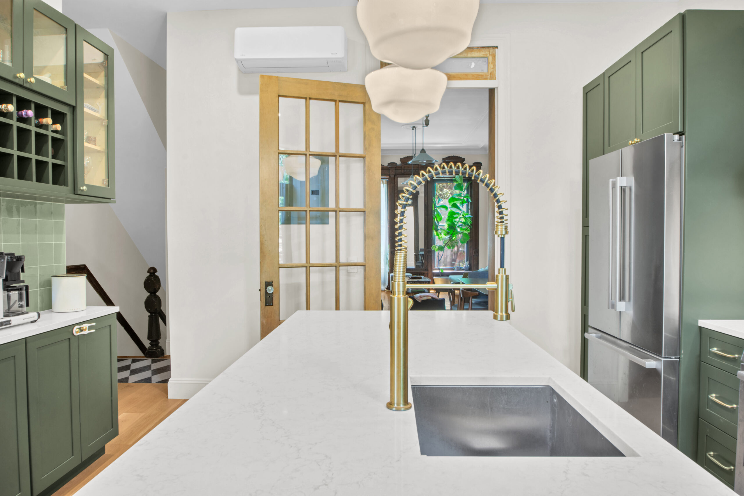 Modern kitchen with green cabinets, white marble countertop island with gold spring coil faucet, stainless steel refrigerator, and wooden door leading to living room.