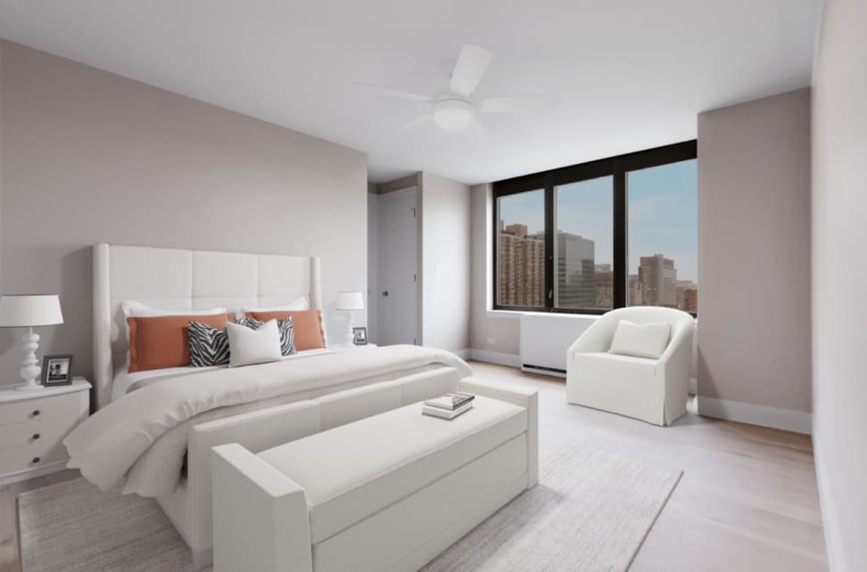 Modern bedroom with white upholstered bed, orange and patterned pillows, white nightstands with lamps, a white bench at the foot of the bed, and a white armchair near a large window showing city buildings.