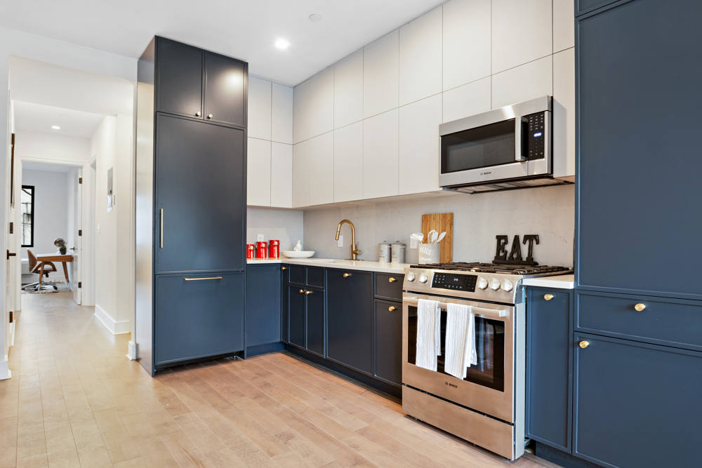 Modern kitchen with navy blue lower cabinets, white upper cabinets, stainless steel stove and microwave, wooden floor, and gold faucet.