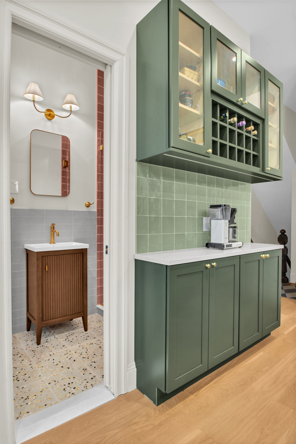 Green kitchen cabinets with glass doors and wine rack, white countertop with coffee machine, next to doorway leading to bathroom with wooden vanity and gold fixtures.