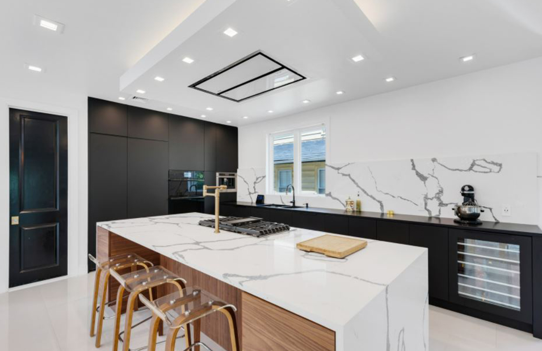 Modern kitchen with a large white marble island featuring a built-in stove and gold faucet, black cabinetry, clear bar stools, and marble backsplash.