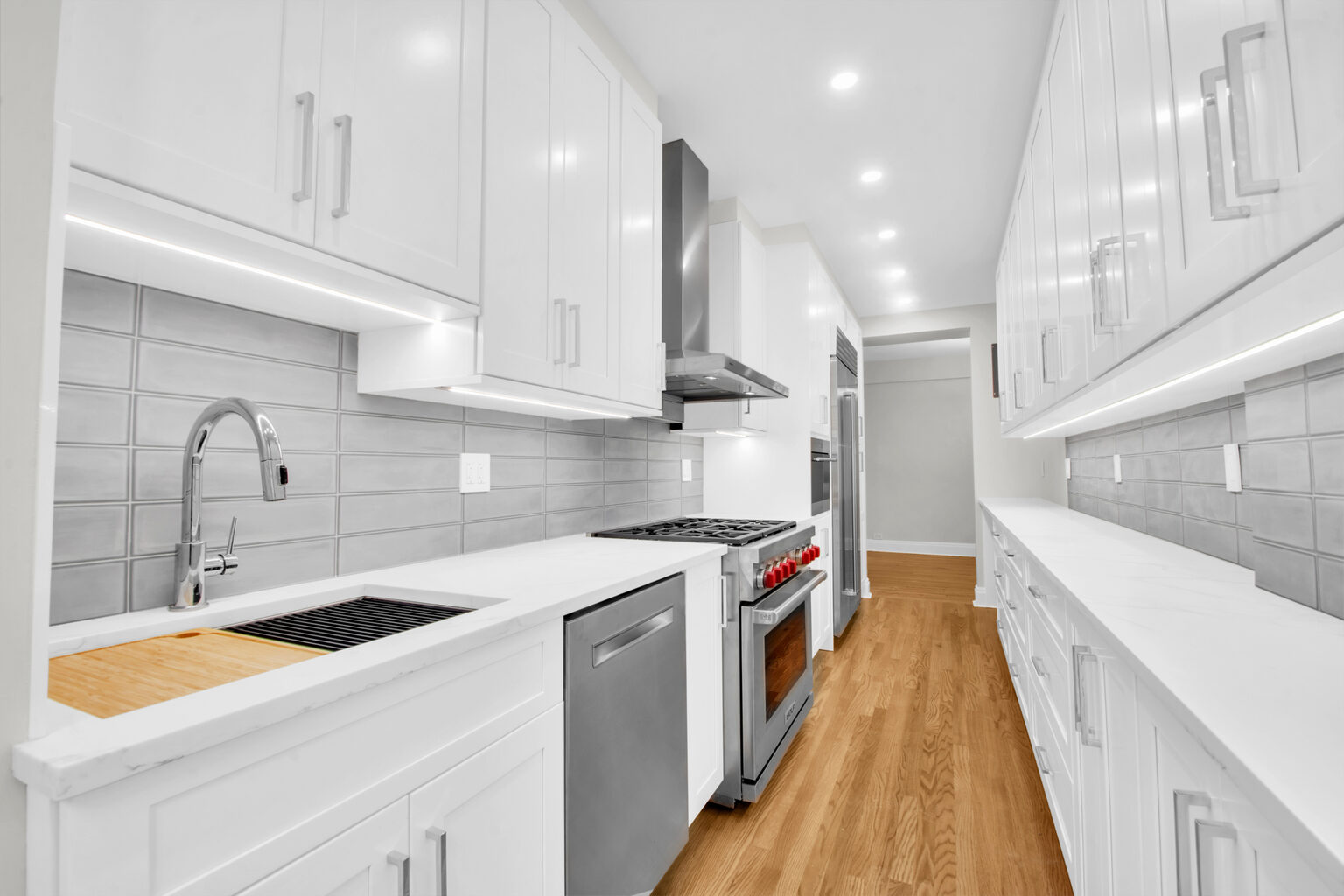 Modern white kitchen with stainless steel appliances, wood floor, and gray subway tile backsplash.