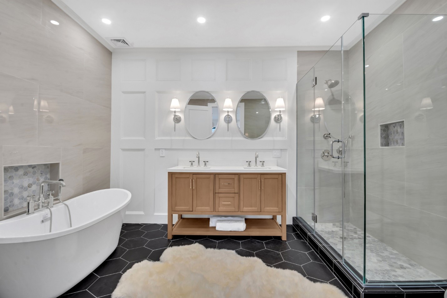 Modern bathroom with black hexagonal floor tiles, white freestanding tub, wooden double vanity with oval mirrors, and glass shower enclosure.