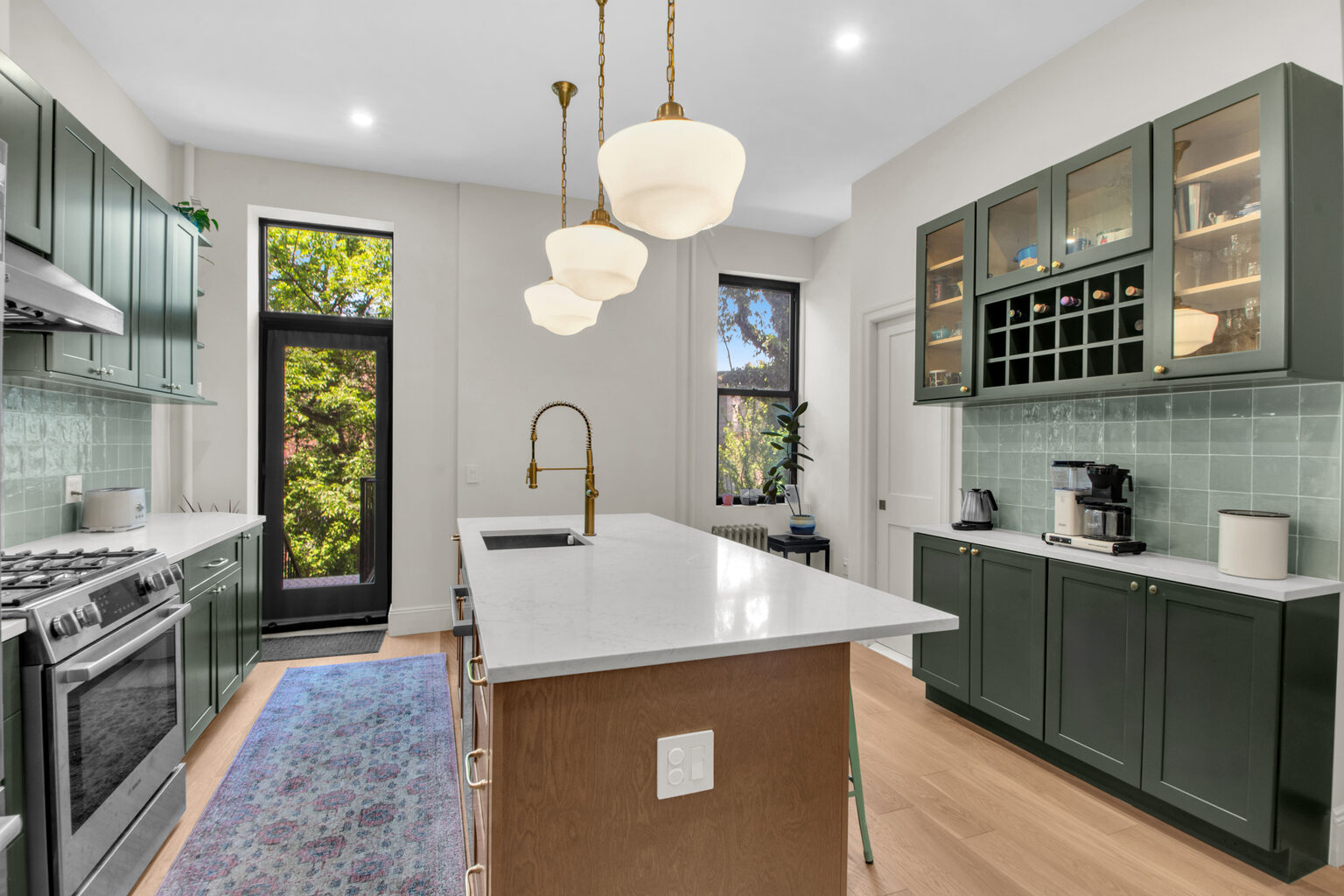 Modern kitchen with green cabinets, white marble island, brass faucet, pendant lights, and a windowed door opening to greenery.
