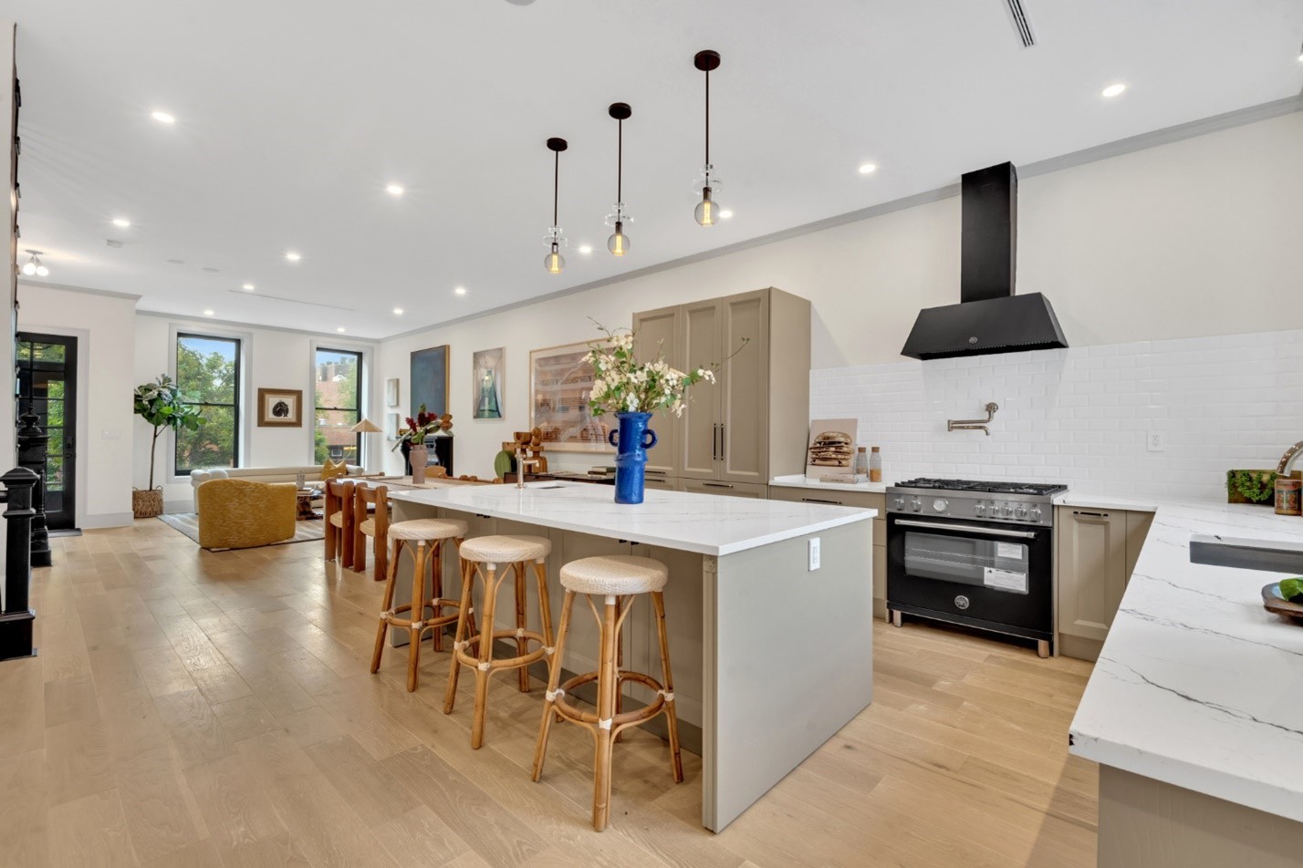 Modern open-concept kitchen and living area with a large island, four wooden stools, black stove with hood, and light wood flooring.