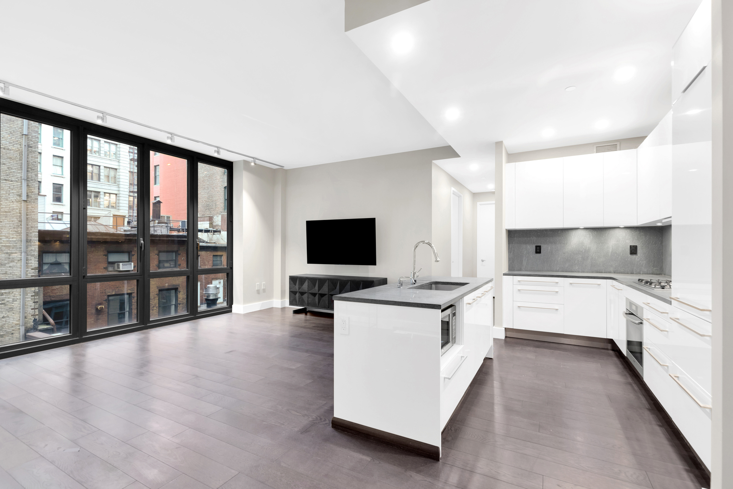 Modern open-plan kitchen and living area with dark hardwood floors, large floor-to-ceiling windows, white cabinets, and a kitchen island with gray countertop.