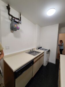 Small kitchen with beige countertop, stainless steel sink, dishwasher, white gas stove, and white walls with exposed gas meter and piping.