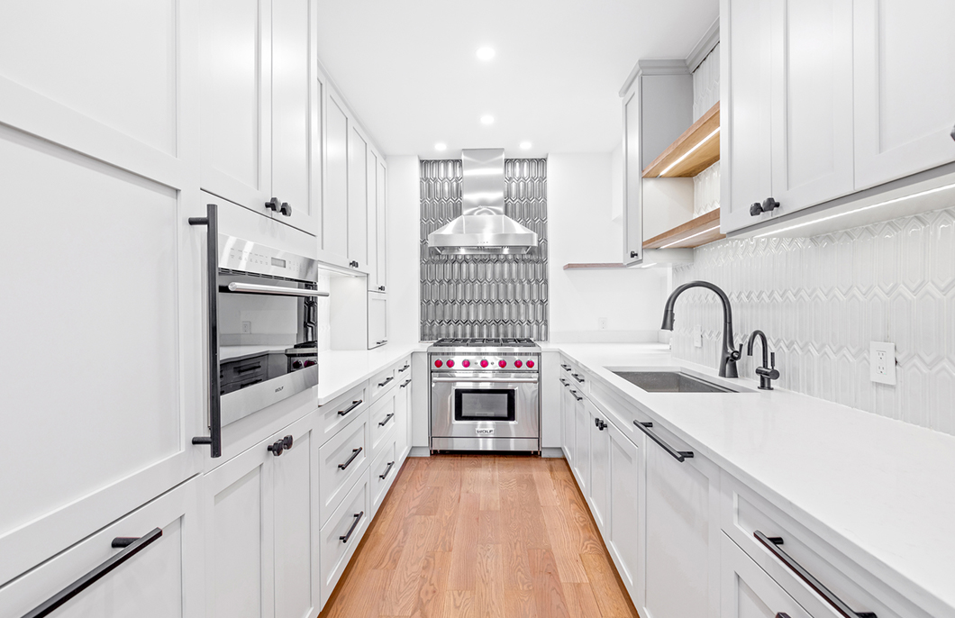 Modern galley kitchen with white cabinetry, stainless steel stove with a stainless steel range hood, white countertops, black hardware, patterned gray tile backsplash behind the stove, and hardwood flooring.