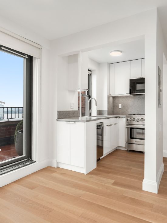 Compact modern kitchen with white cabinets, stainless steel appliances, and light wood flooring, next to a sliding glass door leading to a balcony.