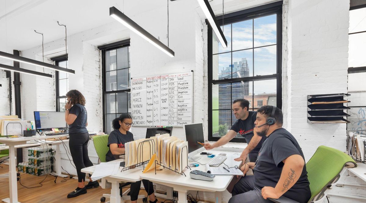 Four office workers collaborating at desks in a bright modern workspace with large windows and a whiteboard displaying a schedule.