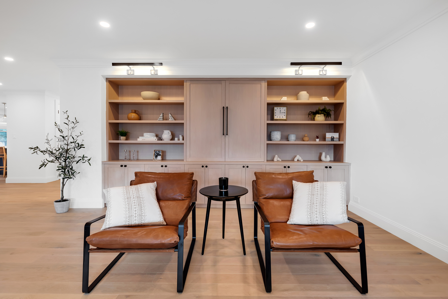 Modern living room with two brown leather armchairs, white patterned cushions, black side table, potted plant, and wooden built-in shelves with decorative items.