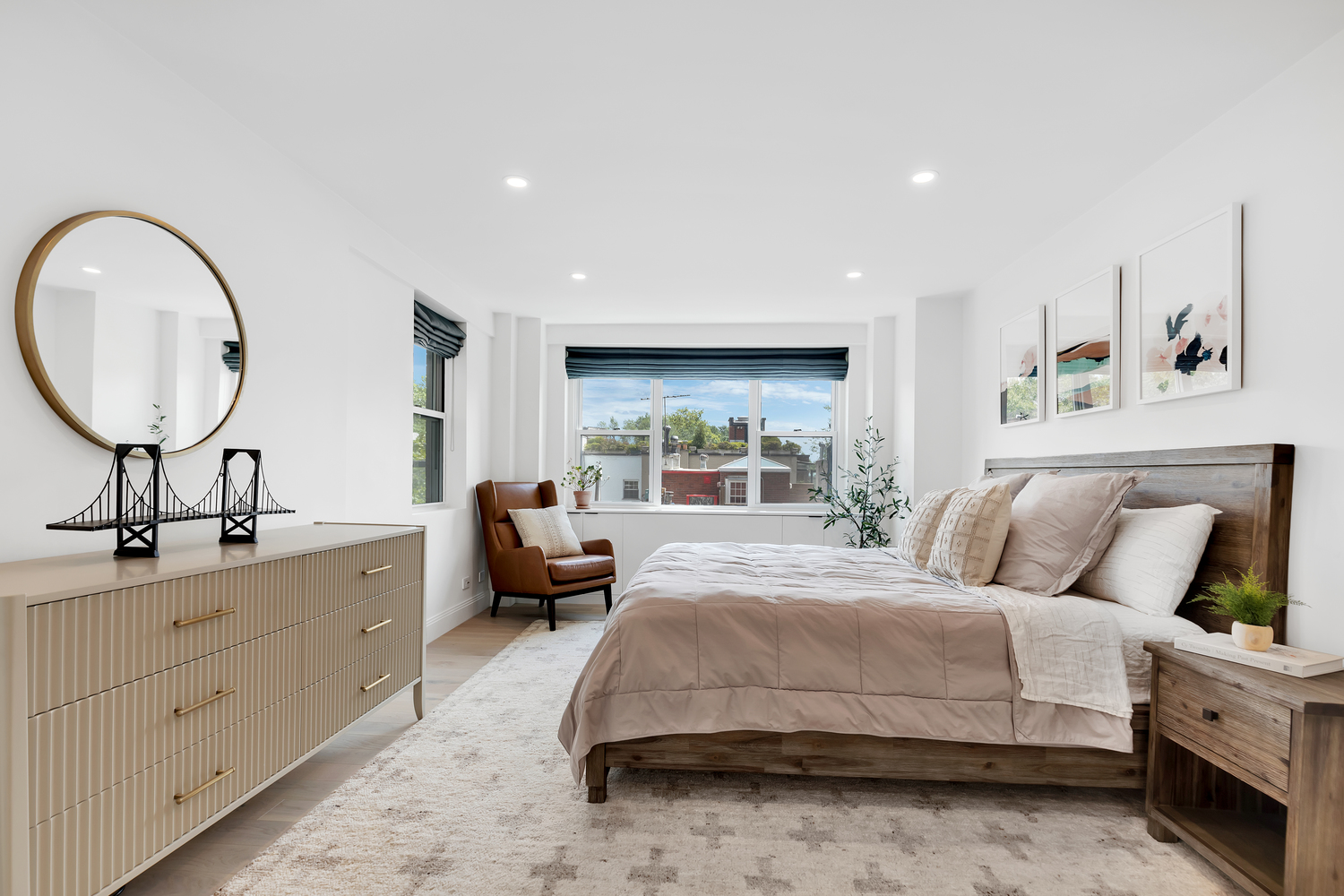 Bright bedroom with a large bed, beige bedding, wooden headboard, three abstract paintings above, and a brown leather chair near a window.