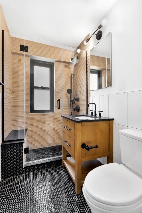 Modern bathroom with black hexagonal tiled floor, wooden vanity with black countertop, glass shower enclosure, and white toilet.
